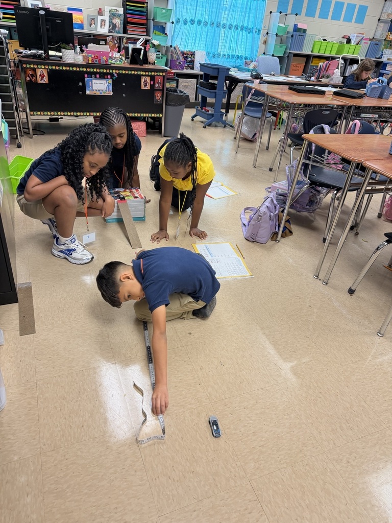 Four children are on the floor in a classroom, engaging in an activity. They are surrounded by tables and chairs.