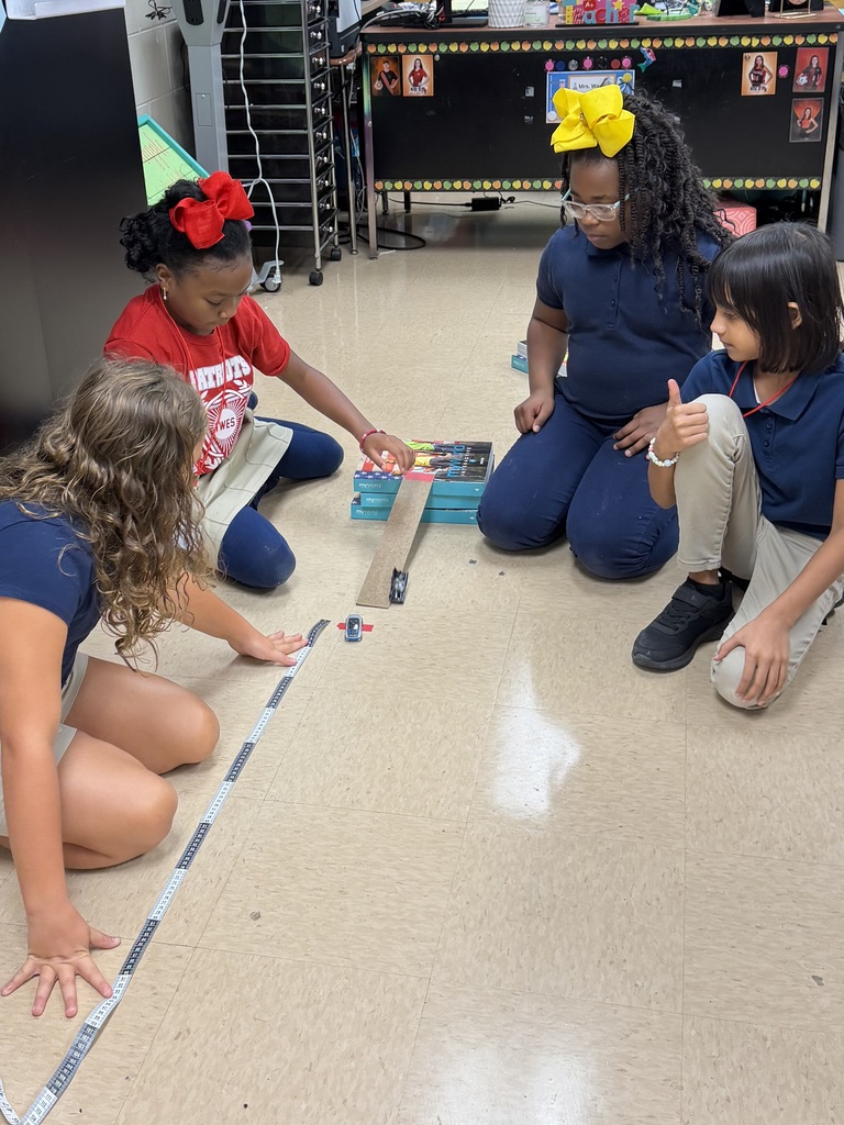 Four young girls are seated on the floor, engaged in a classroom activity. Two adults oversee them, with objects on the floor.