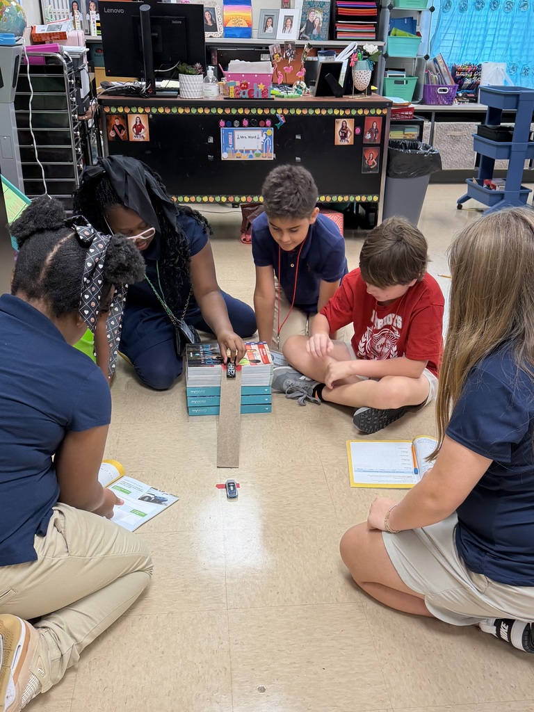 Five children and a teacher sit in a classroom, engaged in an activity with a small robot on the floor.