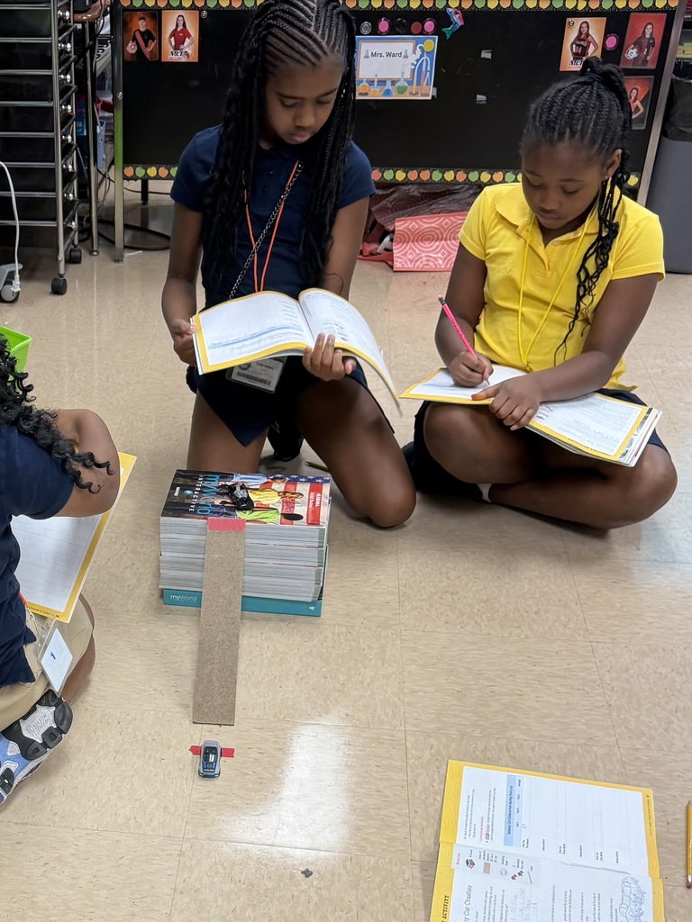 Four children in a classroom setting, two seated on the floor, reading books, with additional books and papers around them.