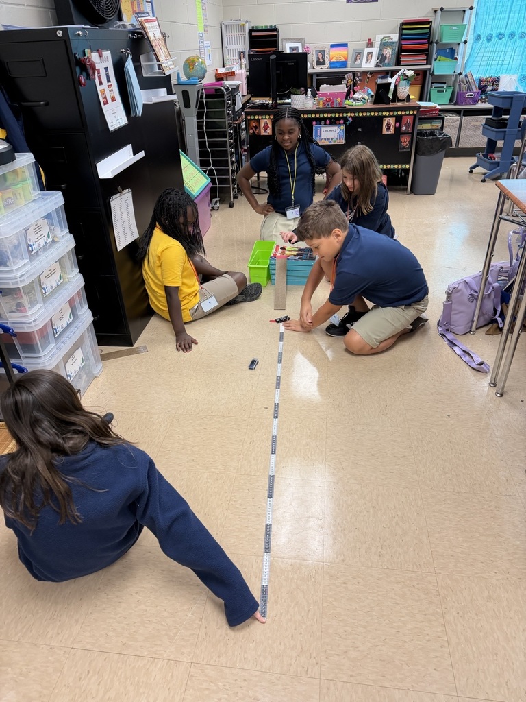 Children seated in a classroom, measuring distance using a ruler, with storage boxes and a table in the background.