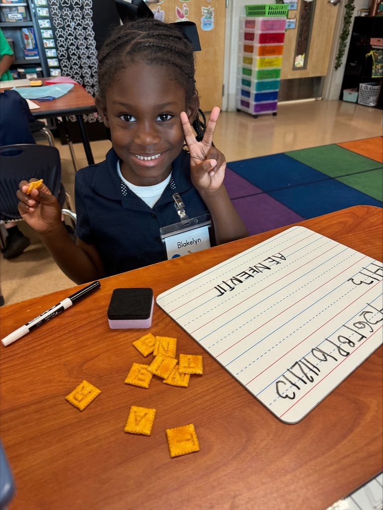 A young girl sits at a table, holding a peace sign, surrounded by yellow tiles and a white board.