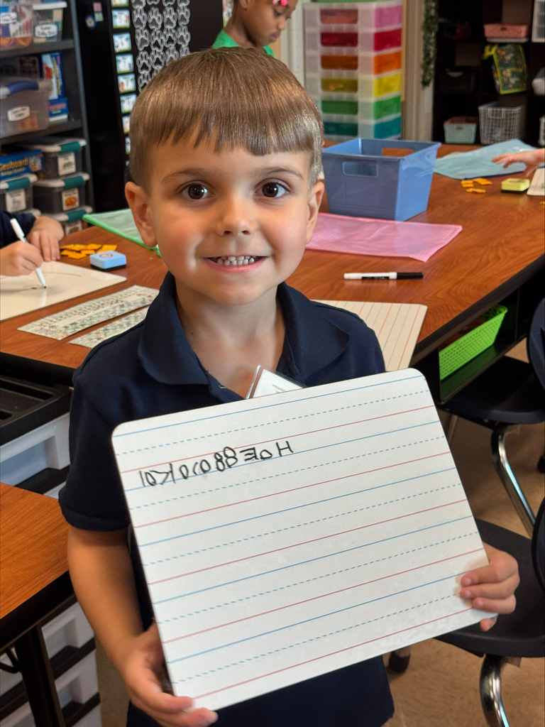 A boy holds a lined paper with "HOMEWORK" written in cursive. He stands in a classroom with other students and desks.