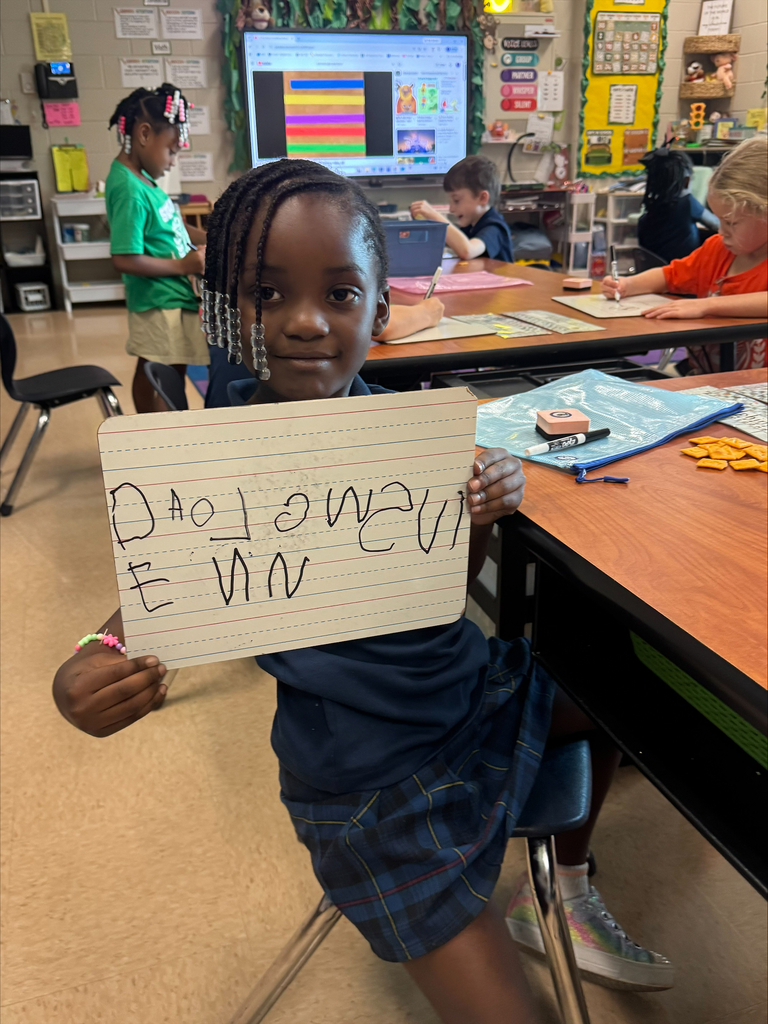A girl in a blue uniform holds a paper with "I am" written on it, smiling in a classroom.