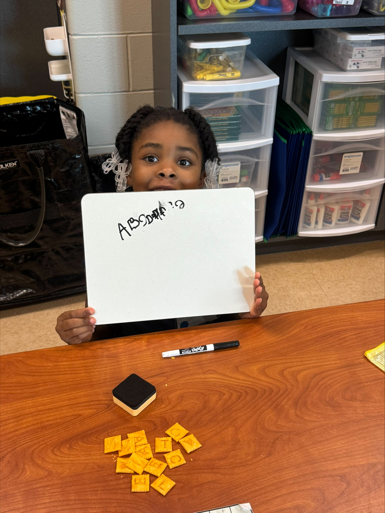 A child holds a whiteboard with "ABSOLUTE 19" written on it, surrounded by yellow tiles. Behind the child is a desk with a black marker and sponge.