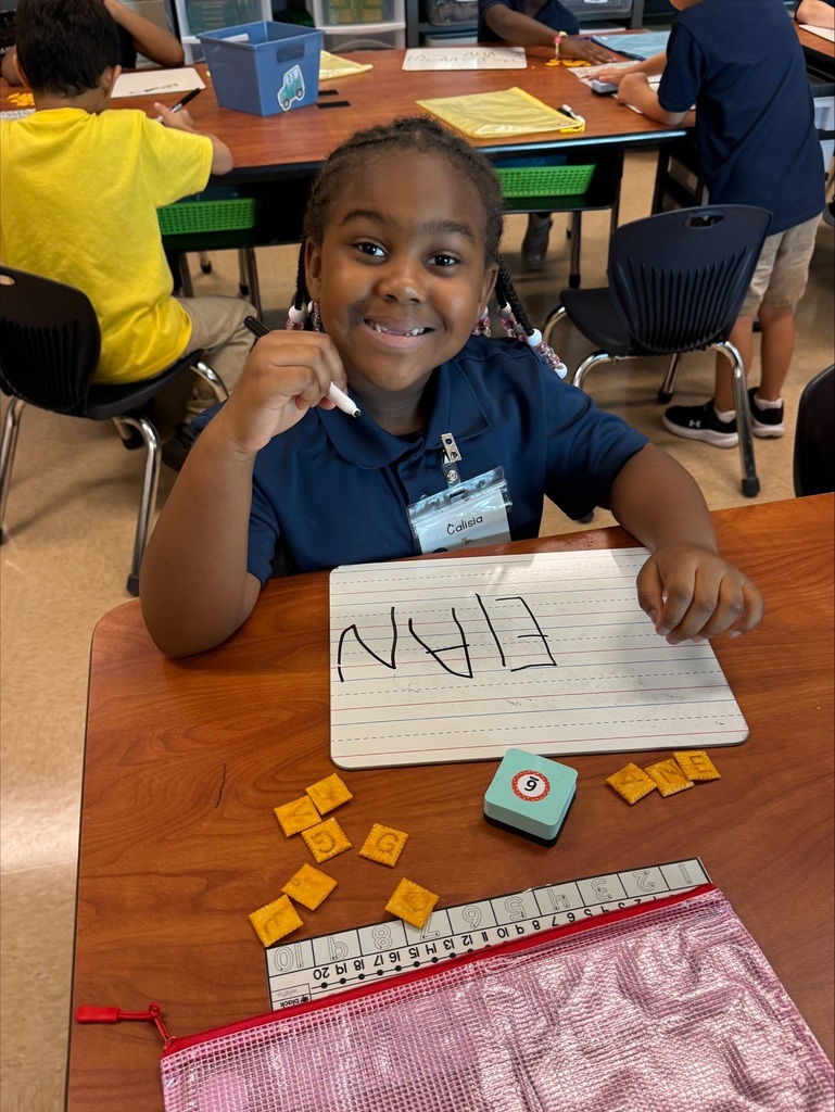 A young boy writes in a notebook, surrounded by letter tiles and a stamper. Other students sit at desks behind him.