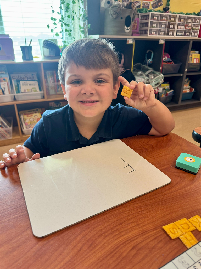 A young boy holds a tile and smiles, seated at a table with a white board displaying numbers.