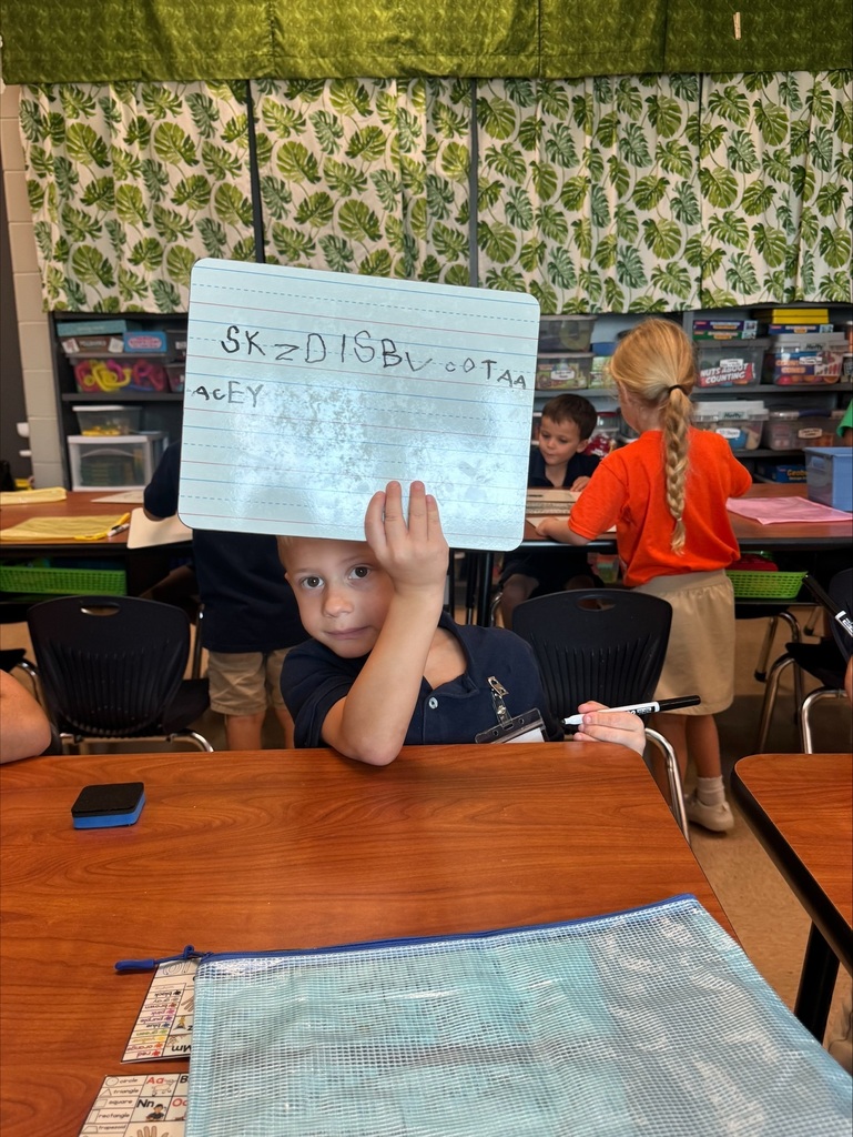 A child holds a whiteboard displaying writing above their head at a desk in a classroom.