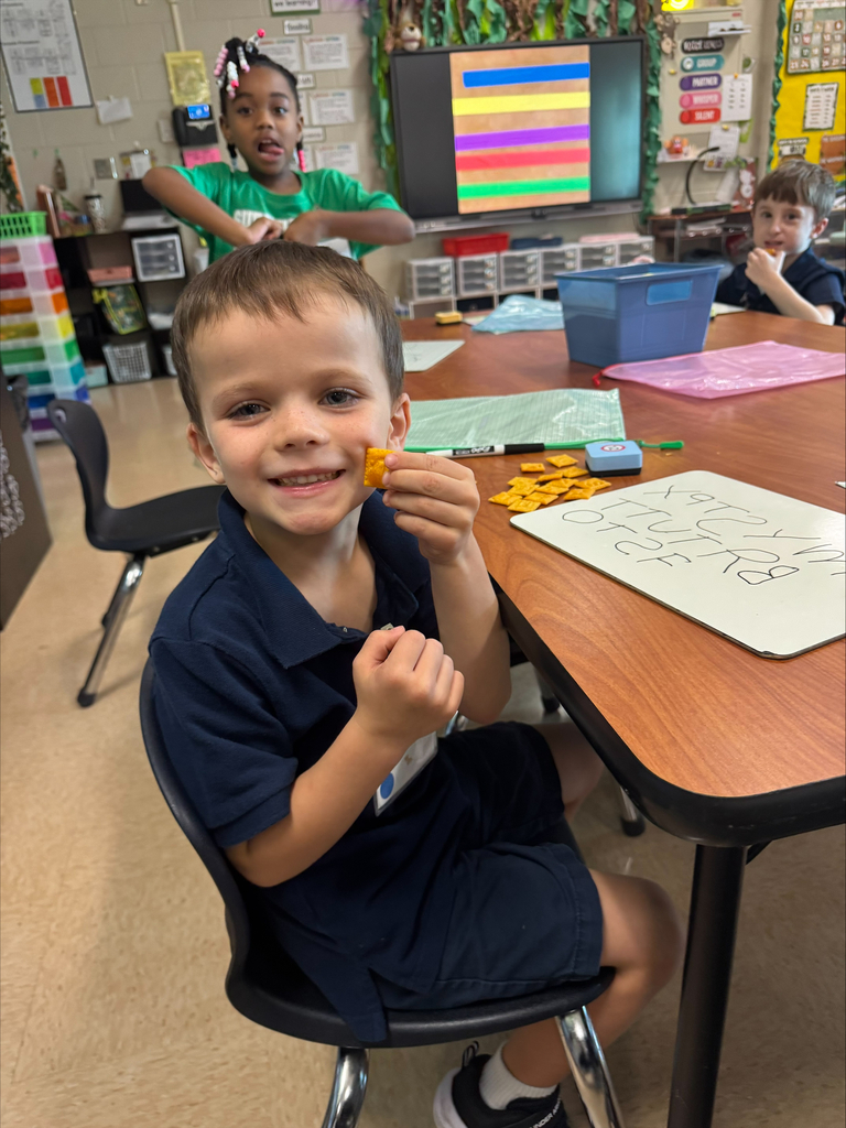 A child wearing a blue polo smiles while holding a piece of food in a classroom.