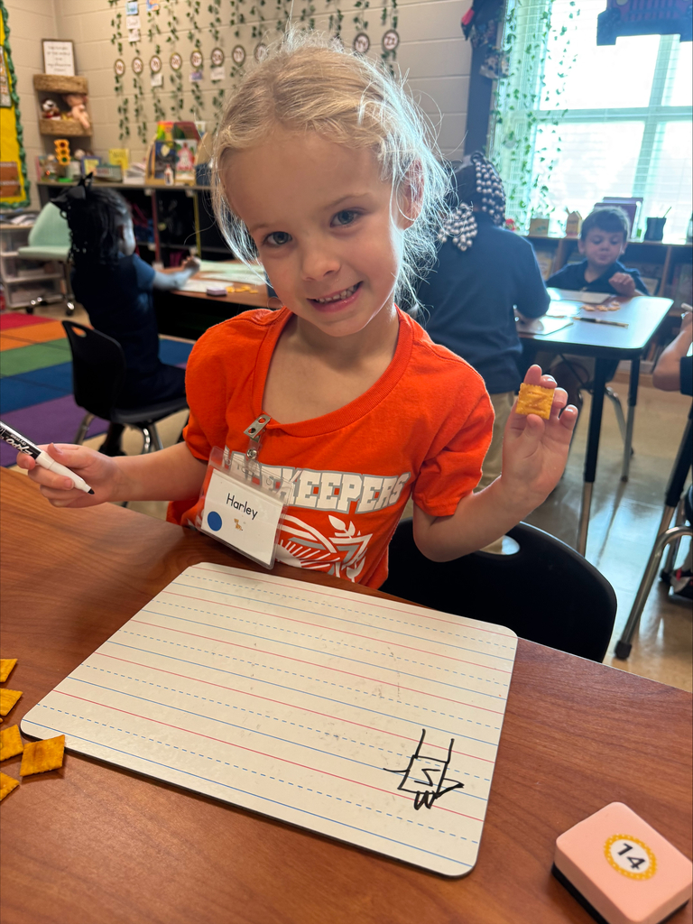 A child in an orange shirt holds a marker and a small object while sitting at a desk with a paper pad.