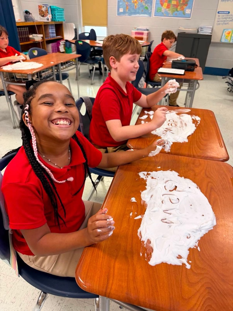 Mrs. Allen’s student writing spelling words in shaving cream on their desk in the classroom. 