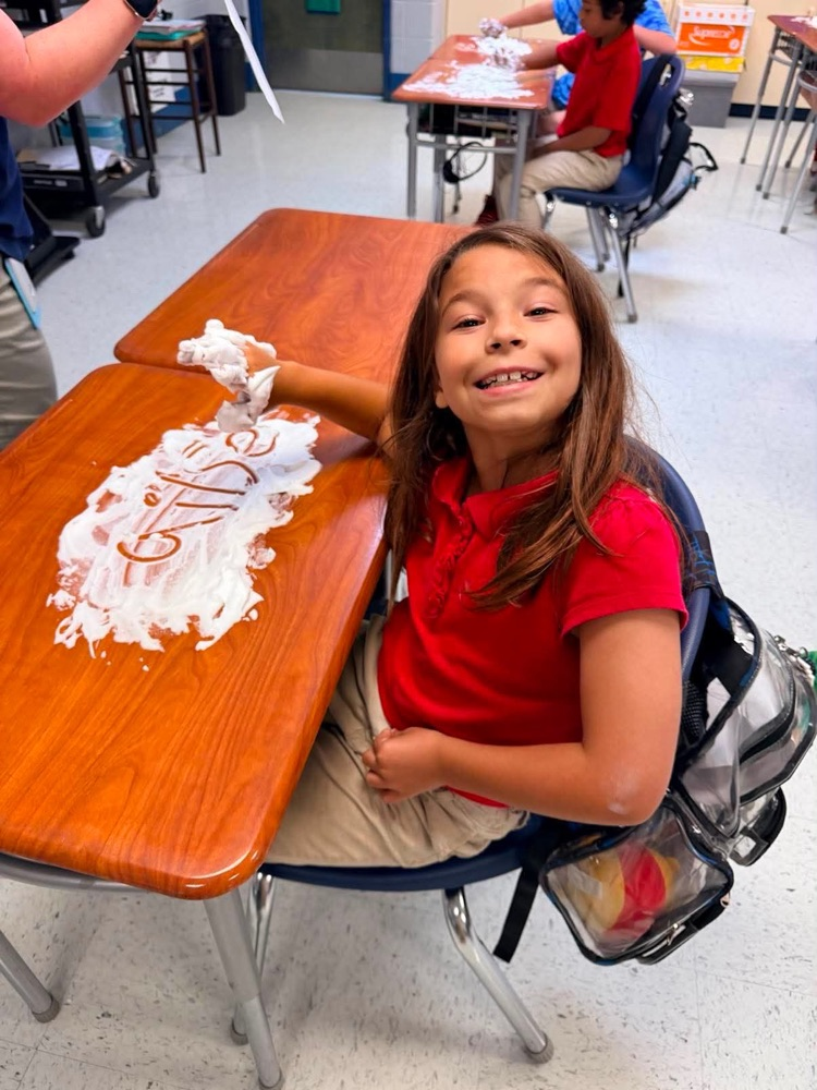Mrs. Allen’s student writing spelling words in shaving cream on their desk in the classroom. 