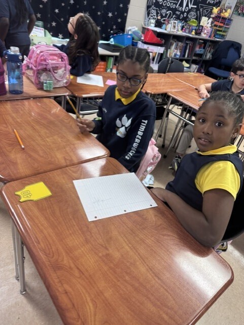 Three students sit at desks in a classroom with bookshelves and a black wall in the background.
