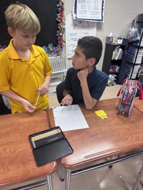 Two boys in school uniforms are seated at a desk. One holds a pencil while the other looks at a paper.