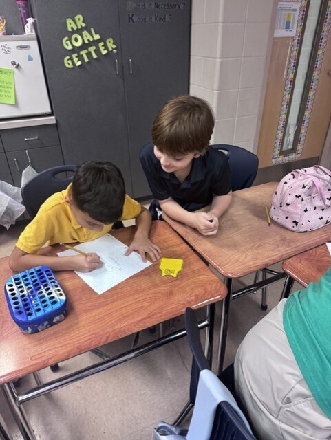 Two boys in yellow and black shirts at desks, writing on paper, with a blue box on the left.
