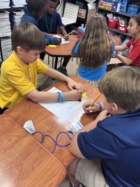 Children in a classroom with desks, one writing while another looks on, and shelves with items.