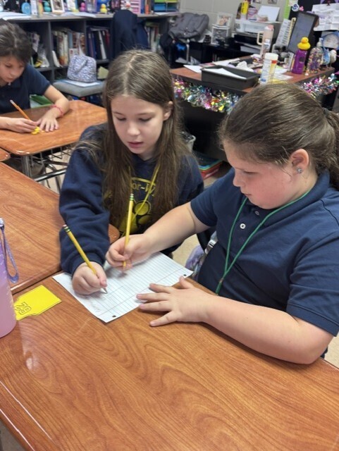 Two students work on paper at desks, one holding a pencil. Another student sits at a desk behind them.