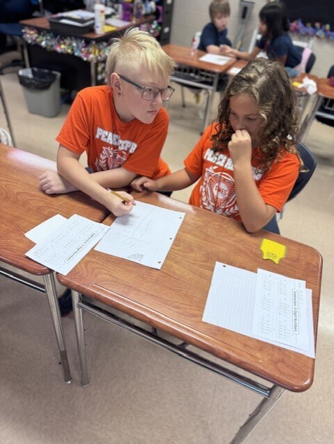 Two students, one with glasses, are seated at a desk, focused on a worksheet. Another student sits at a desk in the background.