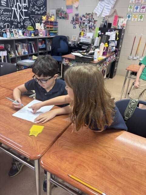 Two students sit at desks in a classroom, one wearing glasses, and they are writing on paper.