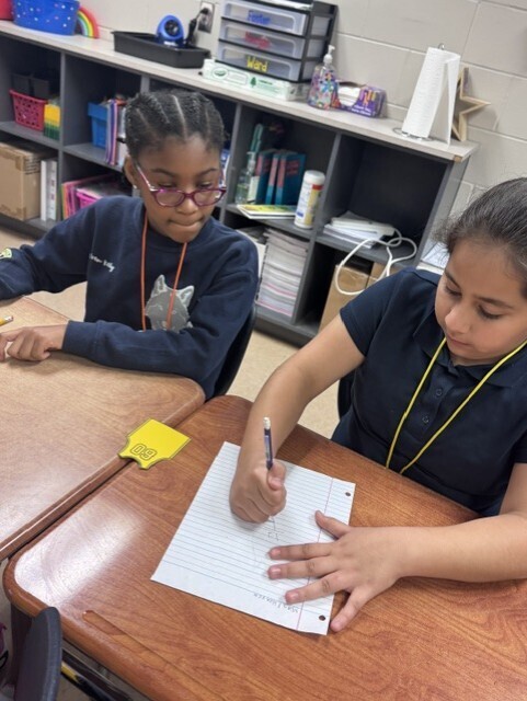 Two students in school uniforms are seated at desks, one writing on paper with a pen.