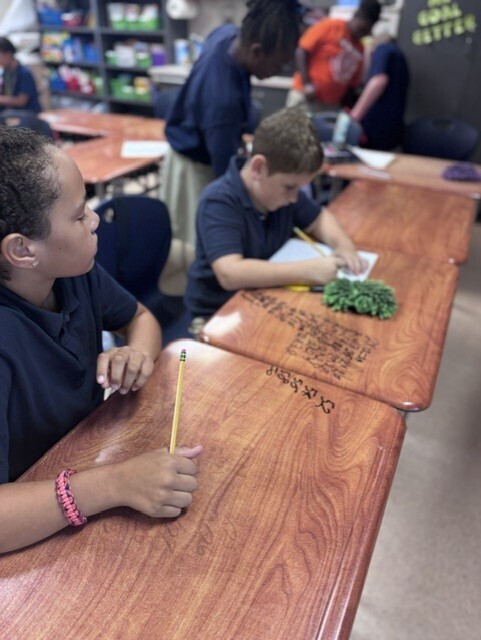 Two students in blue shirts and bracelets sit at desks in a classroom. One has a pencil and paper.