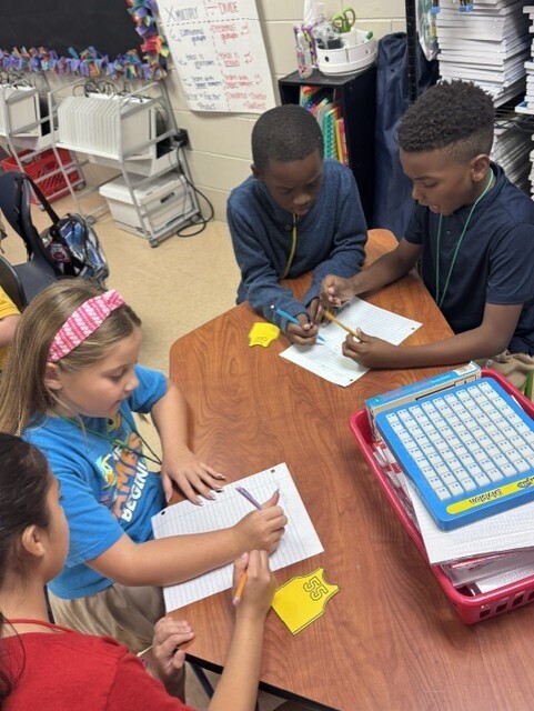 Four children in a classroom setting working at a table. Papers, pencils, and a calculator are on the table.