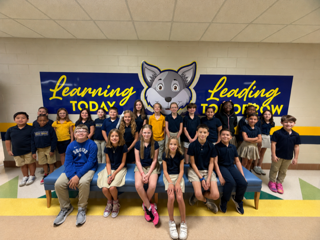 A group of children and adults wearing uniforms sit on a bench in a hallway with blue banners.