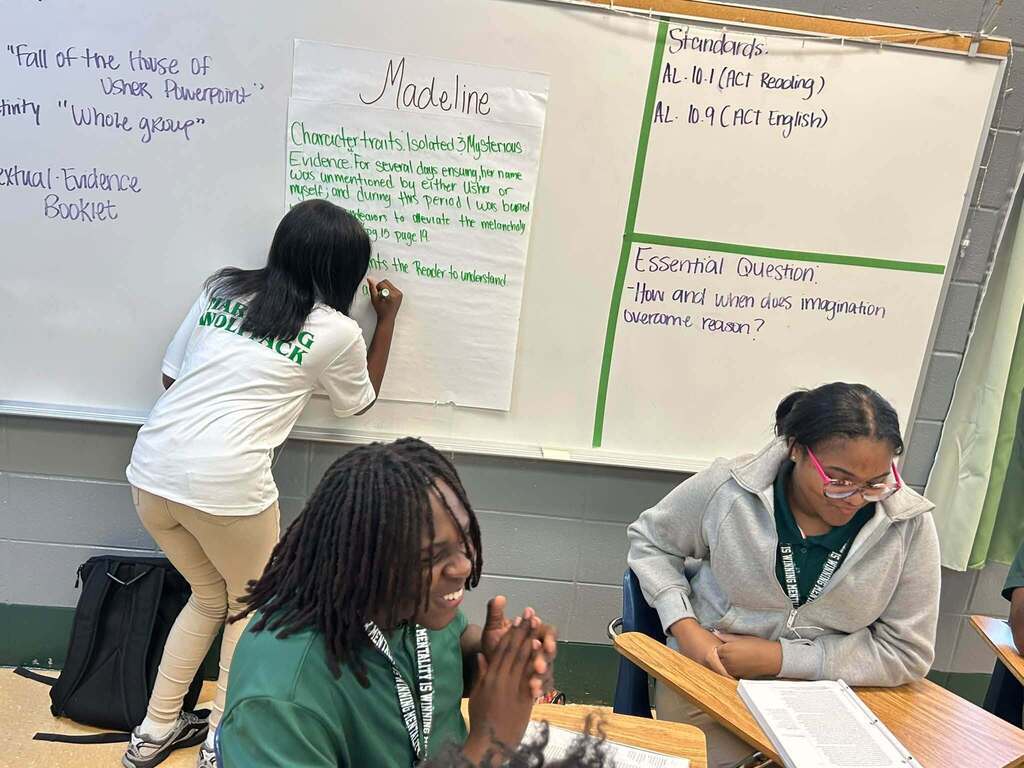 A girl writes on a large poster labeled "Madeline" while two other students sit at their desks