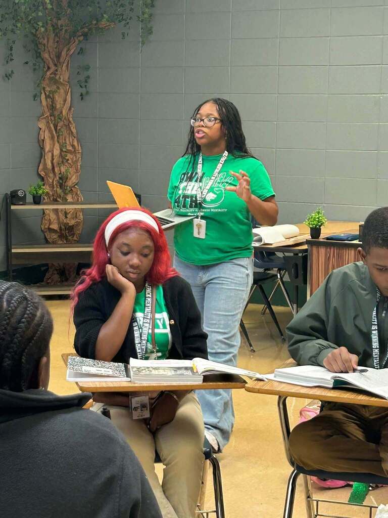 A girl holding a laptop speaks while other students sit at desks
