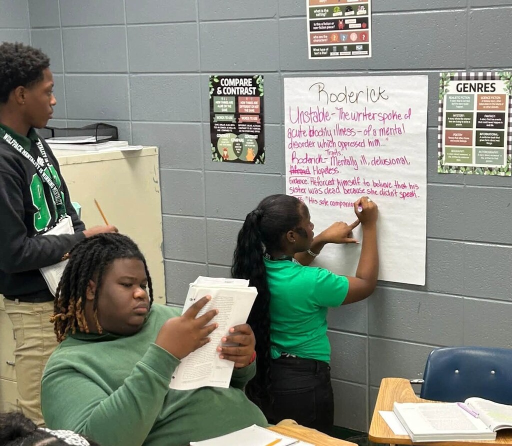 A girl writes on a poster labeled "Roderick" while another student looks at papers at his desk