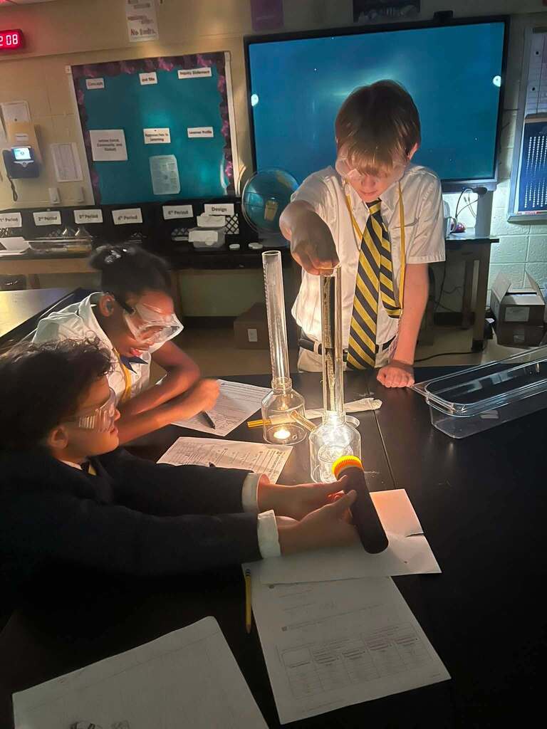 A student pours liquid into a beaker as another student shines a flashlight on it