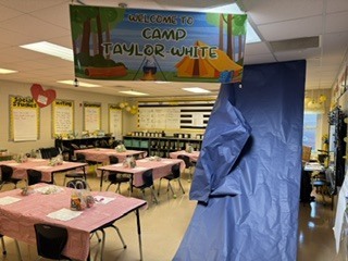 A classroom with tables and chairs. A blue tarp and a banner saying "Welcome to Camp Taylor White" are above.