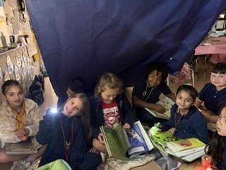 Children sit in a classroom, some under a blue sheet, reading books. A few hold flashlights.