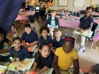 Group of children in blue uniforms sit on floor, holding books and flashlights. Some wear necklaces.