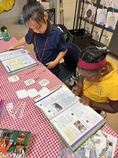 Two children, one in a blue shirt and the other in a yellow shirt, are seated at a table, studying a book.