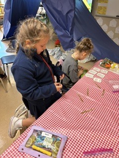 Two young girls seated at a red and white checked table, one holding a needle and thread.