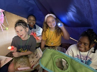 Four kids sit in a tent with flashlights. They smile and appear to be excited.