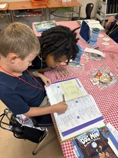Two children sit at a table, focused on a book. One wears a blue shirt and holds a pen.