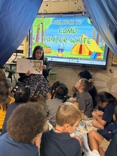 A classroom with a blue curtain backdrop. An adult reads to a group of students seated on the floor.
