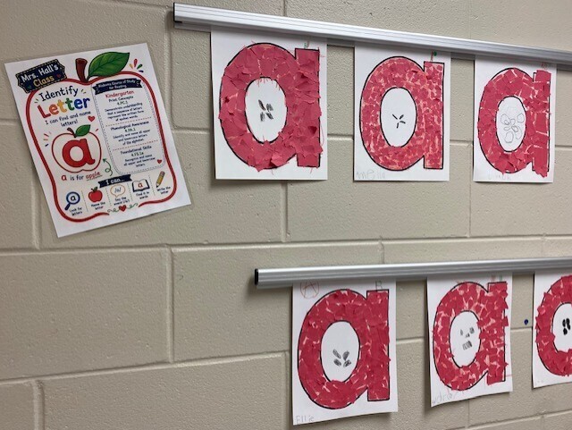 A classroom displays letter banners on a wall with "aaa" and "a" in red. A white-backgrounded paper with red apple graphics and text is pinned to the left.