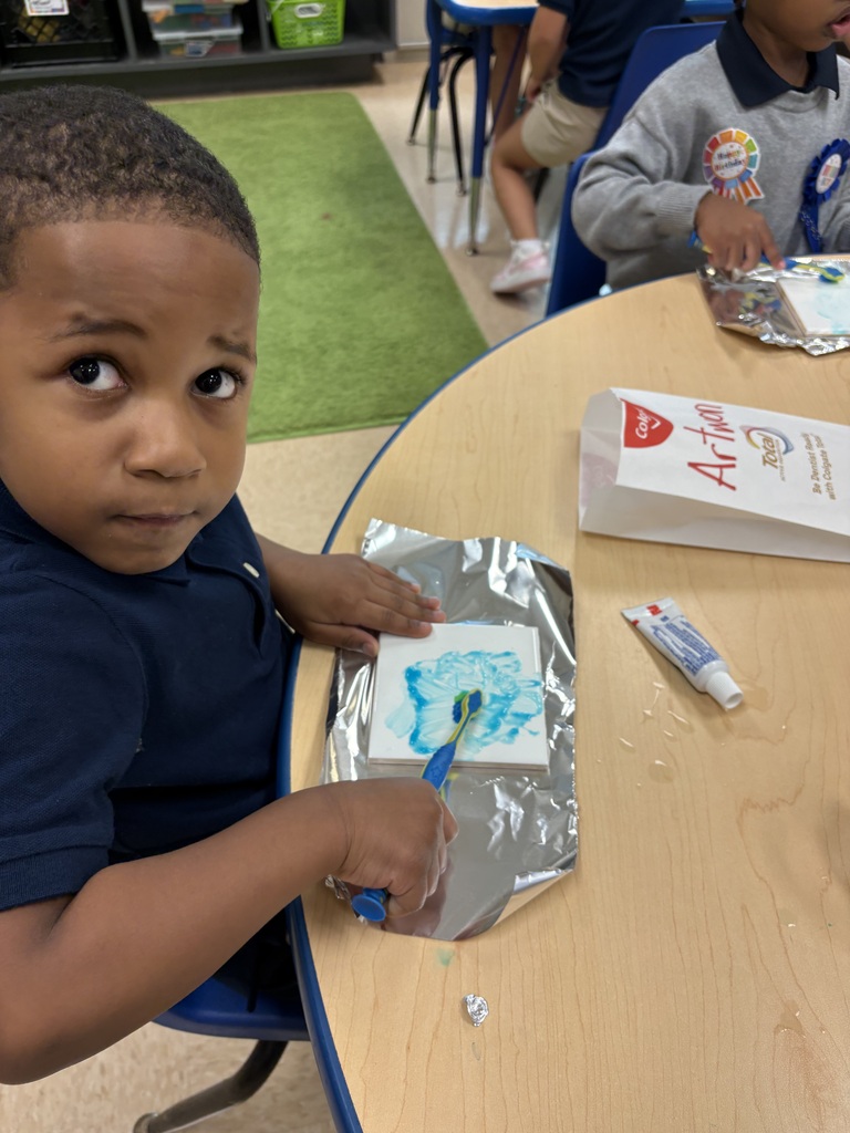 A young child in a blue shirt paints a picture on a table, with a box of Art-E and a toothbrush nearby.
