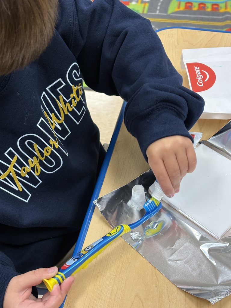 Child in blue sweatshirt holding toothbrush with toothpaste on a desk with a foil-covered container.