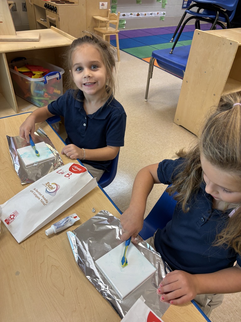 Two young girls in navy blue shirts and khaki pants sit at a table. One holds a paintbrush.