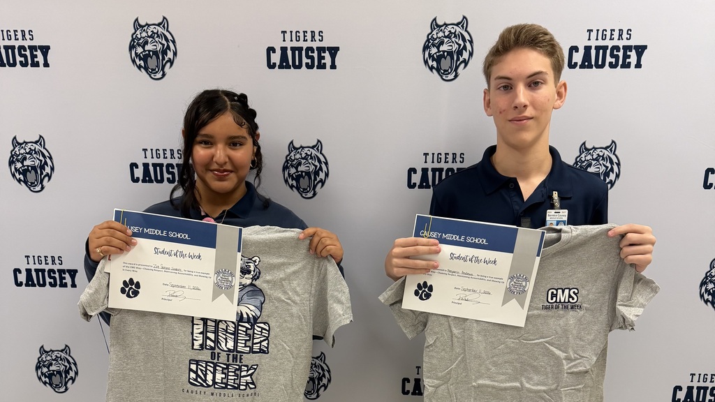 Two young people hold certificates and matching gray shirts in front of a wall with a tiger logo.