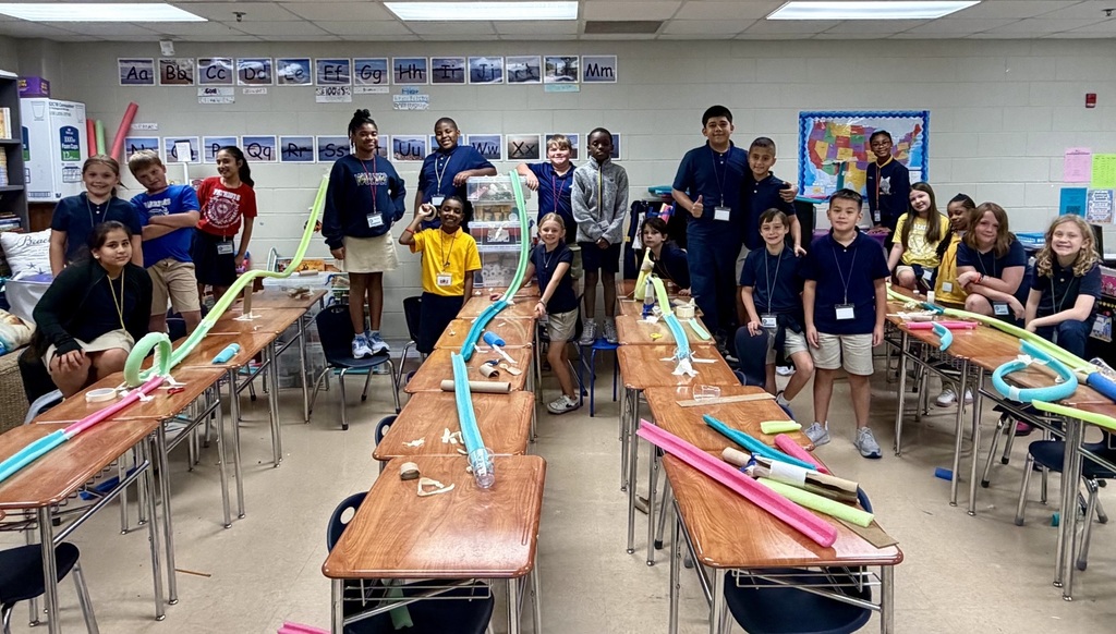 Students in a classroom gather around tables, showcasing various colorful objects, likely part of a school activity.