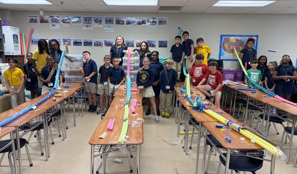 A group of children in a classroom, some standing, others seated, all holding long colorful tubes.