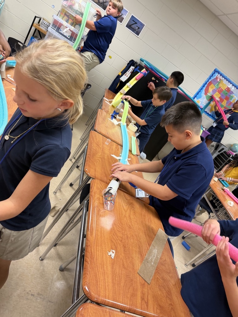 Kids in blue shirts and khaki shorts in a classroom setting, working with colorful materials at desks.