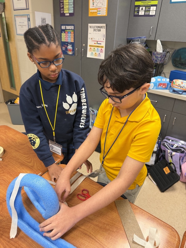 Two children work on a blue craft, one holding scissors. They are in a classroom with a table, cabinets, and a bulletin board.