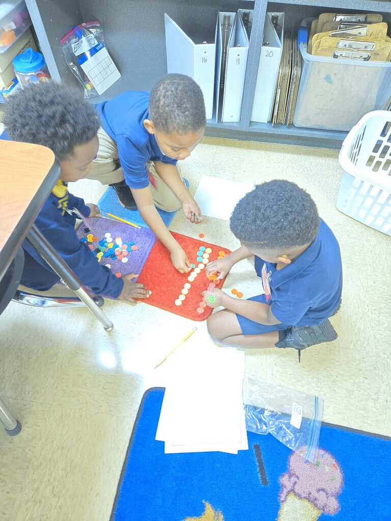 Three children in blue uniforms seated on the floor, one building with blocks and others observing.