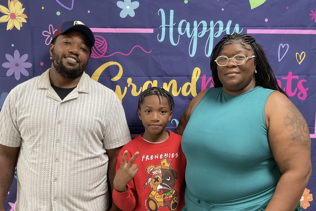 A family of three stands before a backdrop with "Happy Grandparents" written in cursive. A man and woman wear casual clothing.